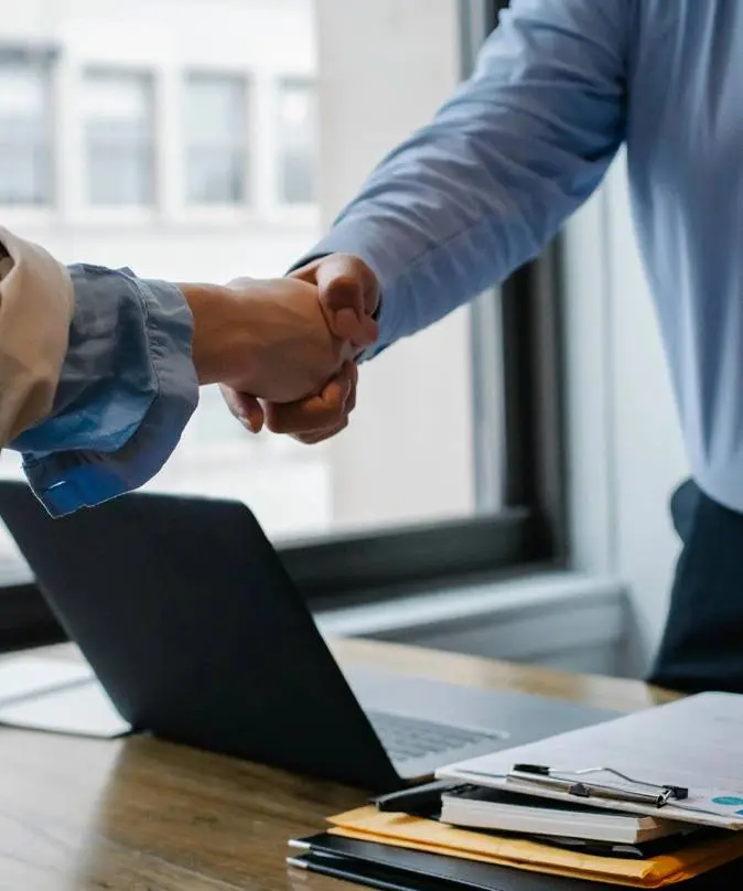 Two people shaking hands over a table with a laptop.